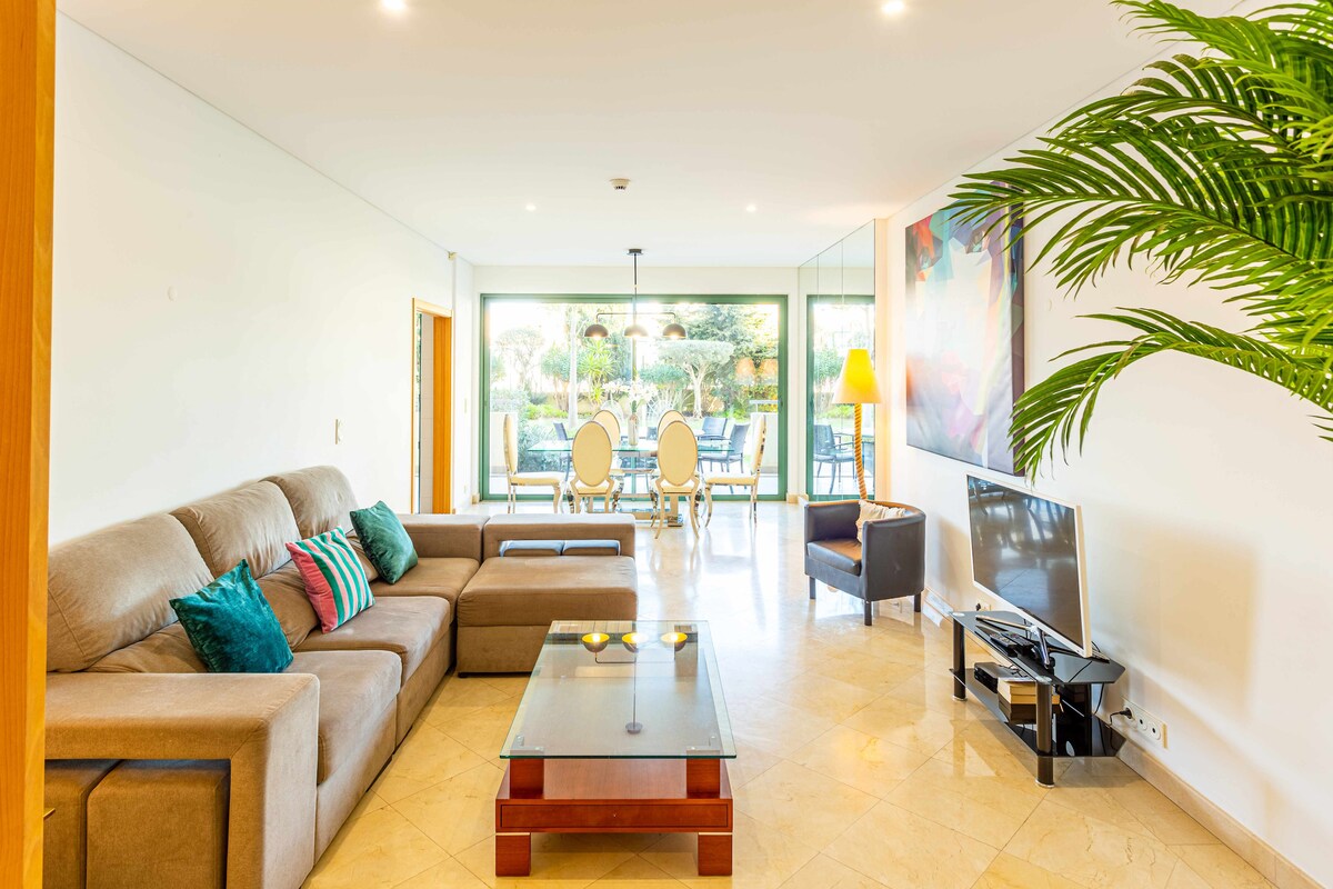 A spacious living area is displayed, featuring a comfortable sectional sofa adorned with various cushions. A glass coffee table is positioned on a tiled floor, reflecting light from large windows overlooking the garden. A flat screen TV sits on a stand, with a dining area visible in the background.