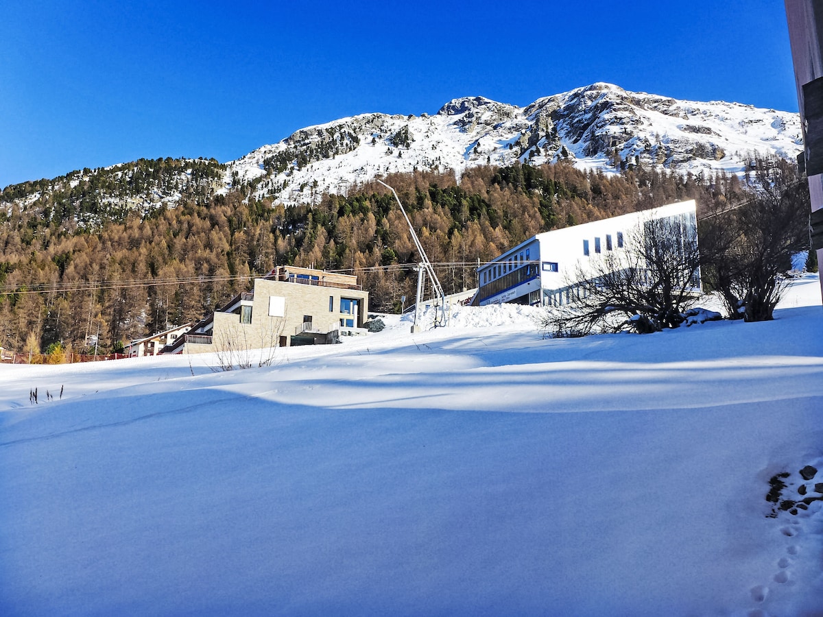 A winter landscape is presented, showcasing a wide expanse of snow covering the ground. The nearby buildings display modern architecture, partially surrounded by trees, with snow-capped mountains in the background under a clear blue sky.