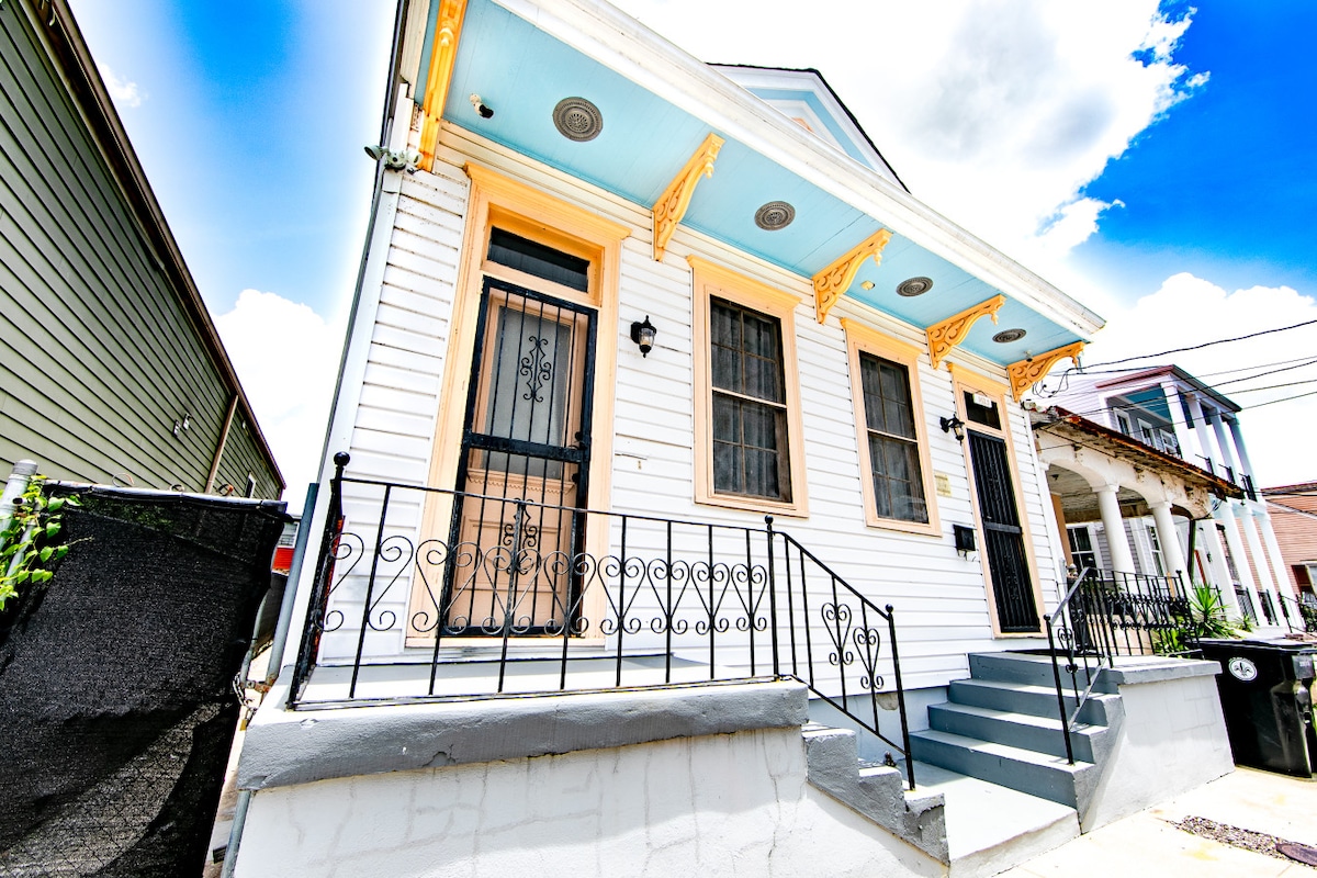 A charming single-family home features a pastel blue ceiling above the entrance. The exterior showcases white siding with ornate yellow trim and a wrought iron railing. Steps lead up to the front door, framed by large windows that allow natural light to enter.