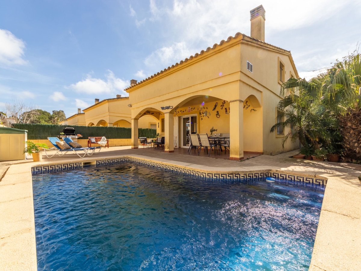 The spacious outdoor area features a private swimming pool framed by a sun deck, with lounge chairs positioned for relaxation. A covered terrace offers dining space, surrounded by lush greenery and sunny skies.