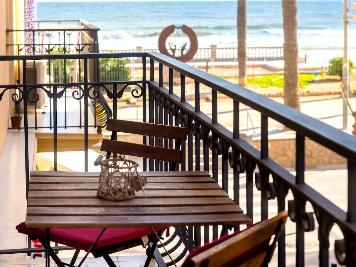 A wooden table with matching chairs is positioned on the balcony, overlooking the sea. The railing features an ornamental design. Soft waves can be seen in the distance, with palm trees lining the promenade, enhancing the coastal view.