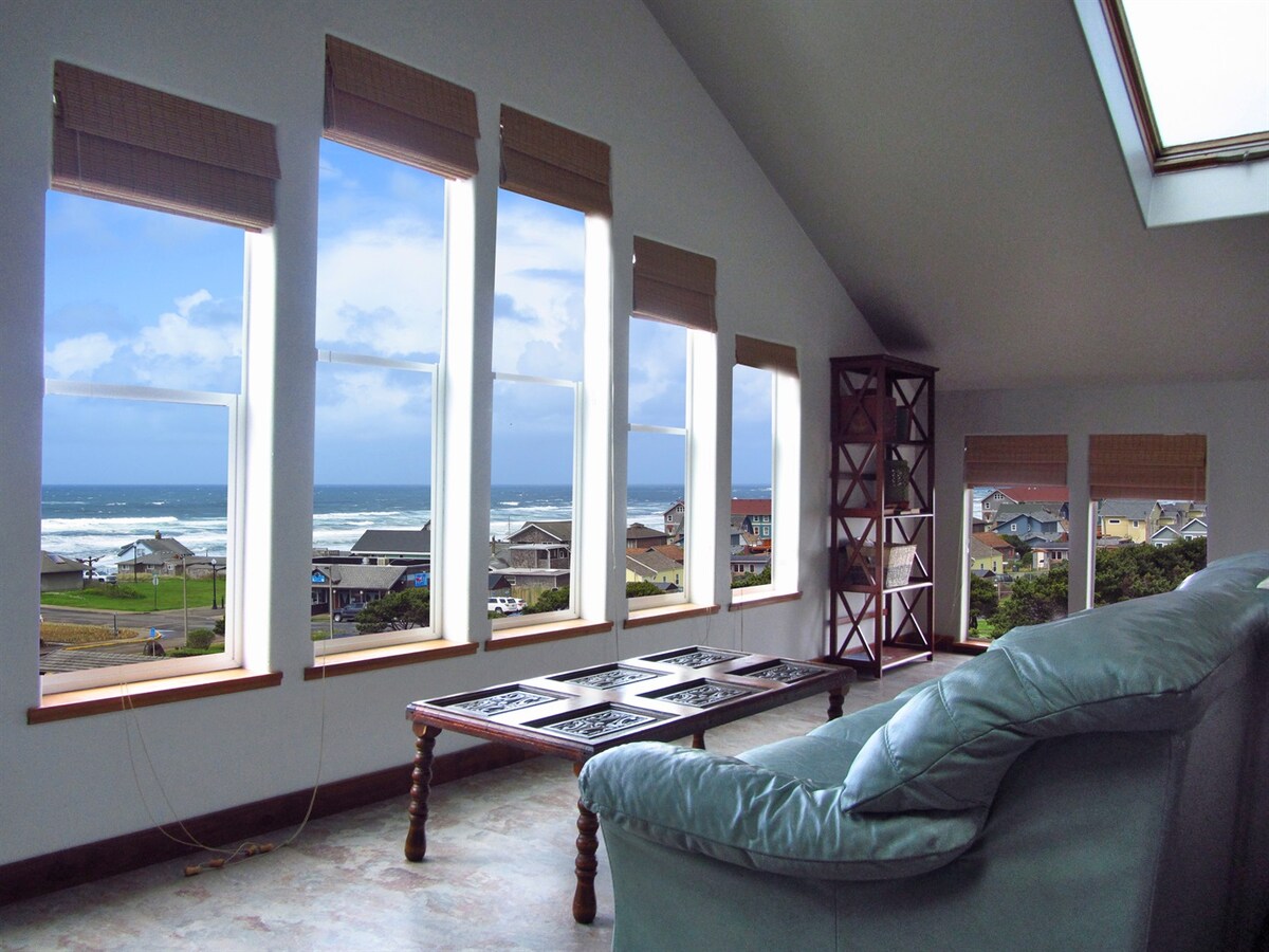 Large windows provide expansive ocean views and natural light, framing the horizon and coastal scenery. A comfortable seating area is arranged with a low table, enhancing the space's openness. Light-colored walls and high ceilings contribute to a bright and airy atmosphere.