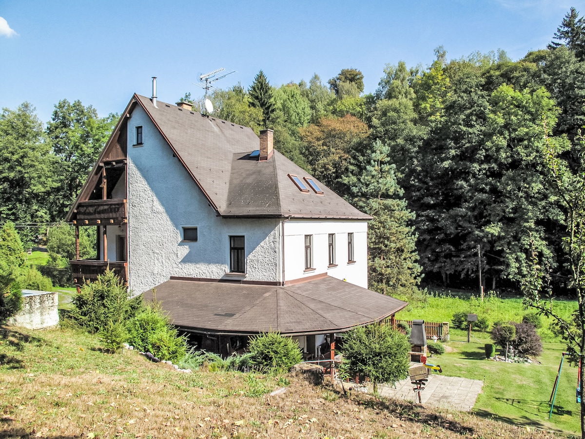 A two-story house is set in a natural landscape, surrounded by trees and greenery. The exterior features a combination of white and dark wood elements, with a sloped roof and several windows. A spacious lawn and garden area are visible, enhancing the serene environment.
