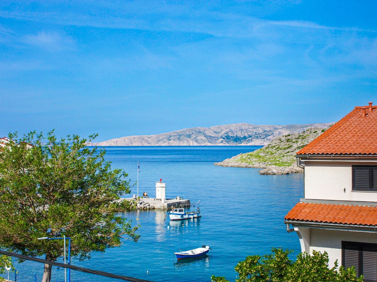A serene view of the sea is captured, featuring a small boat moored near a coastal structure. Lush green trees frame the scene, with a backdrop of rolling hills and a clear blue sky, enhancing the tranquil seaside atmosphere.