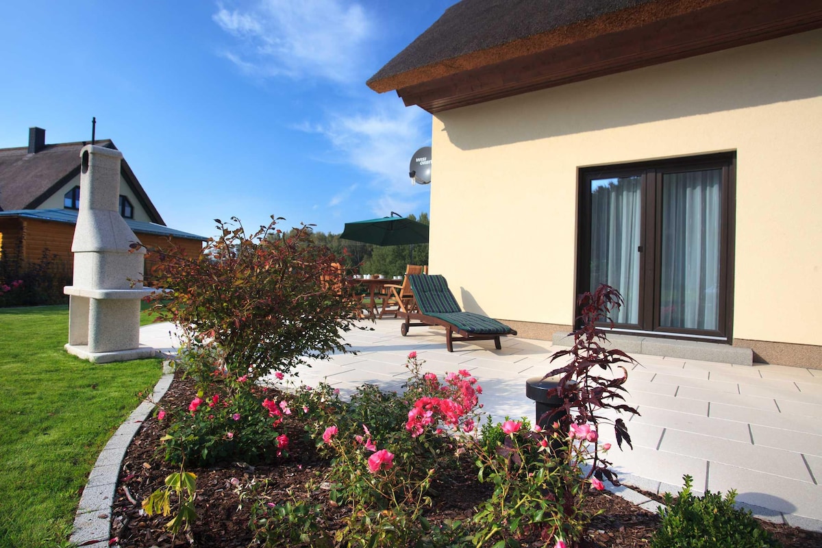 A well-maintained terrace area features a sun lounger and a stone grill, surrounded by colorful flower beds. A dining table with chairs is visible under a green umbrella, providing a pleasant outdoor space for relaxation and meals.