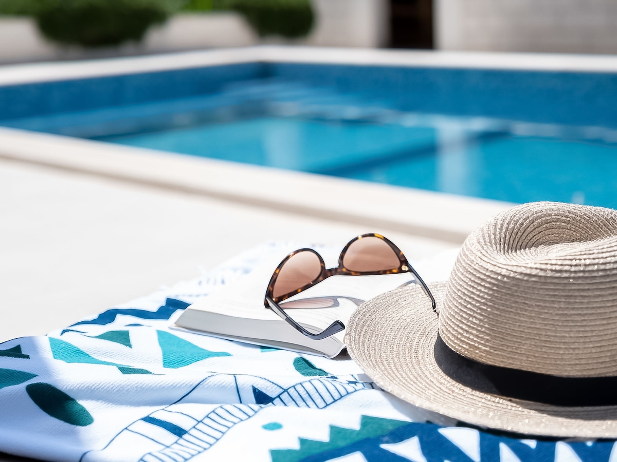 A sun lounger is placed near a shimmering swimming pool, accompanied by a wide-brimmed hat and sunglasses. A book rests on the lounge, suggesting a relaxing atmosphere. The pool's blue water reflects the surrounding area, enhancing the tranquil setting.