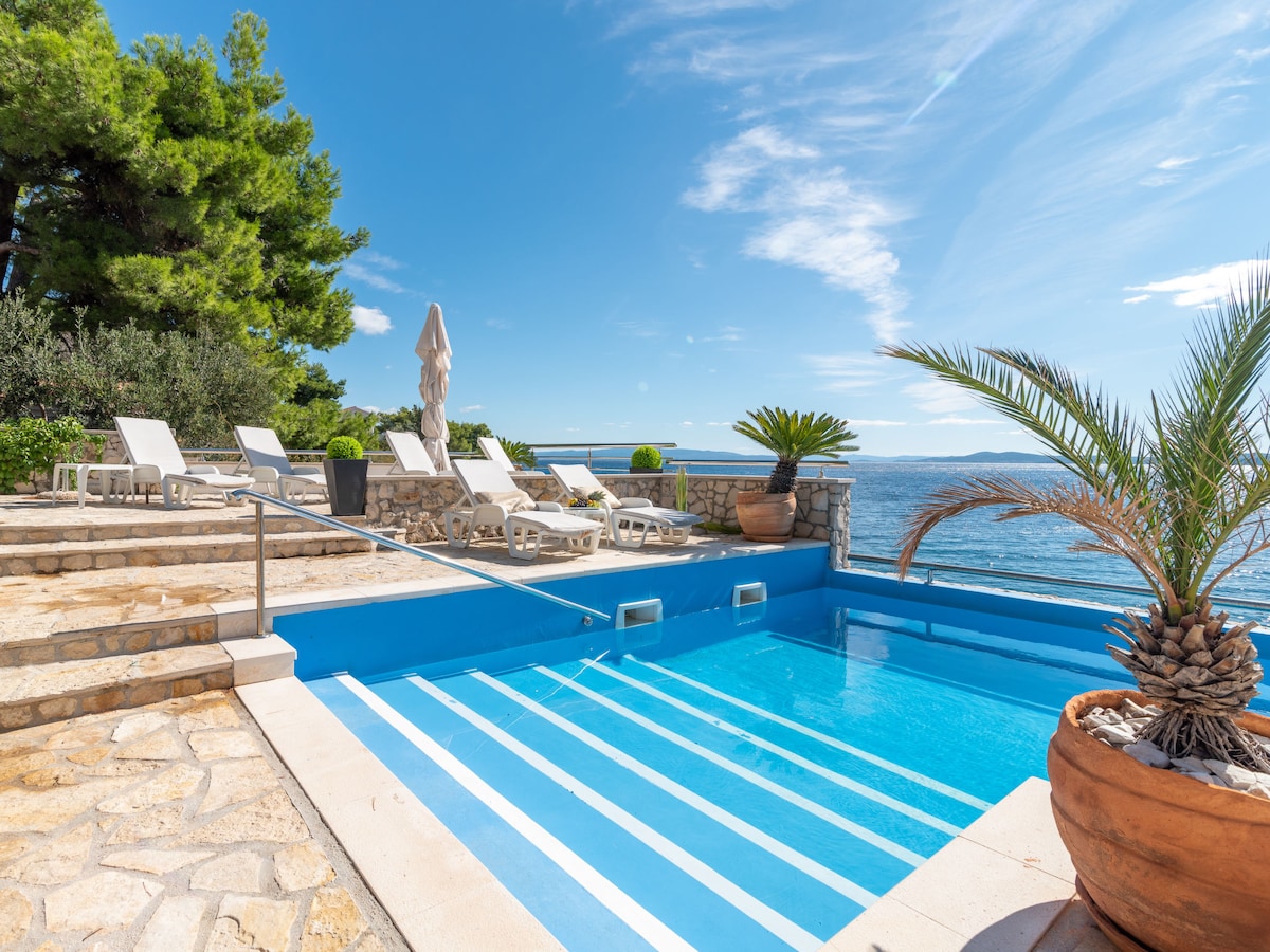 A swimming pool with a turquoise surface is framed by reclining chairs and tropical plants. The surrounding stone patio offers a comfortable area for relaxation. The clear blue sky and sea create a serene backdrop, enhancing the tranquil environment.