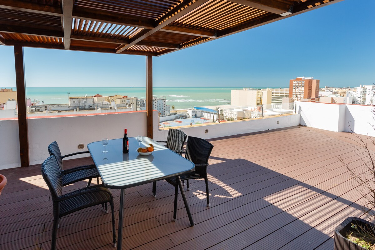 A spacious terrace features a dining table set with chairs, overlooking a panoramic view of the sea. Sunlight filters through a wooden pergola, casting shadows on the deck, while the horizon stretches with distant buildings and clear blue skies.