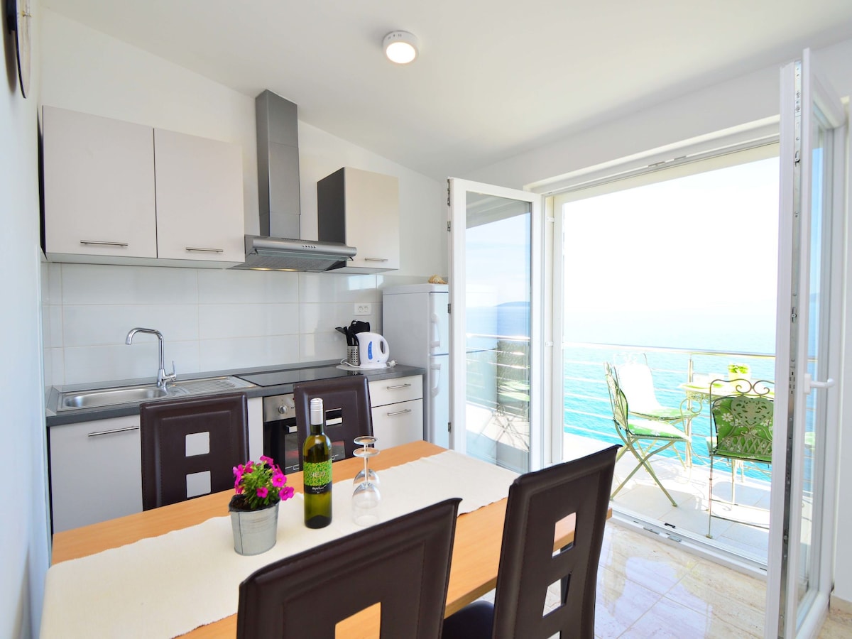 An open kitchen area features modern appliances, including a stovetop and kettle, along with a spacious dining table set for four. Large glass doors lead to a balcony with seating, showcasing a panoramic view of the sea.
