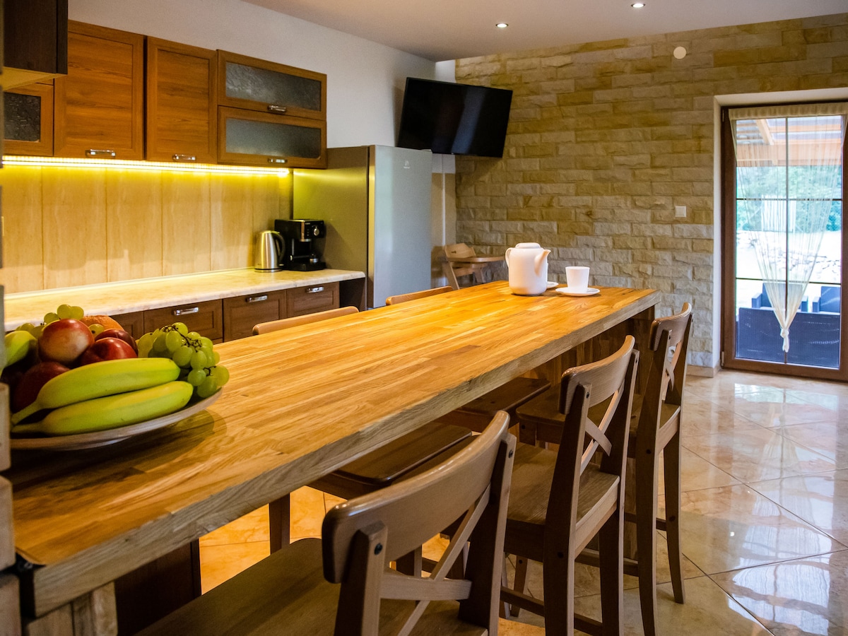 The kitchen features a spacious wooden countertop with seating, complemented by a variety of fruits on a platter. Modern appliances, including a coffee maker and refrigerator, are visible. Stone accents add texture to the walls, while soft lighting enhances the inviting atmosphere.