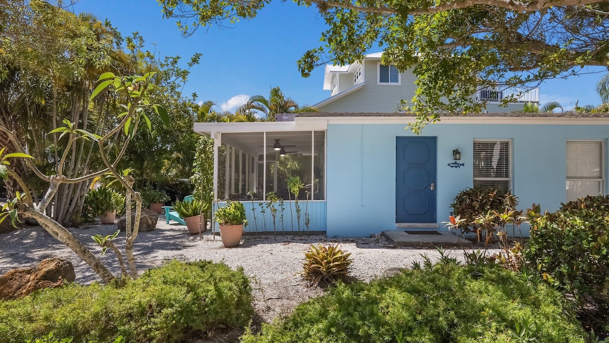 A ground-level duplex is showcased, featuring a soft blue exterior. A screened lanai is visible at the front, surrounded by lush tropical landscaping. Potted plants add greenery to the outdoor space while a clear blue sky complements the charming setting.