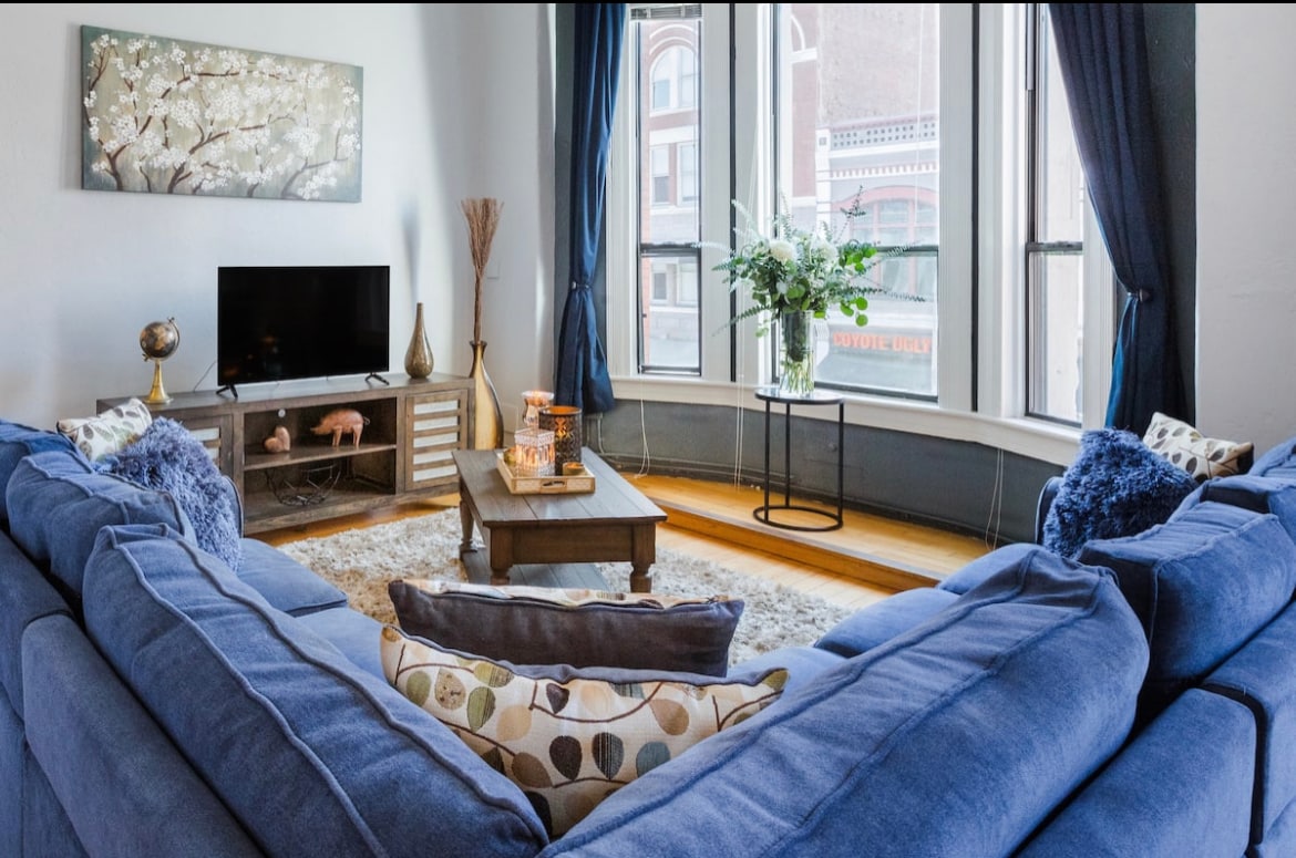 The living room features a large blue sectional sofa arranged around a wooden coffee table. A Smart TV sits on a console, and natural light floods the space through bay windows adorned with dark curtains. Decorative artwork and fresh flowers add subtle accents.