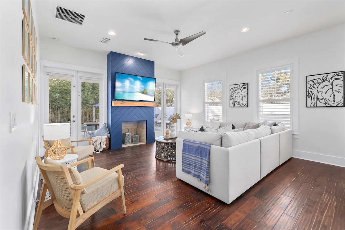 A bright living area features a spacious sectional sofa, complemented by a decorative blue wall. Large windows invite natural light, while a smart TV is mounted above a modern fireplace. Stylish wall art adds character to the room, which is accented by warm hardwood flooring.