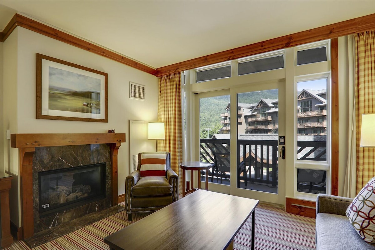 A welcoming living area features a cozy seating arrangement with a chair and sofa positioned around a low coffee table. Large windows allow natural light to fill the space, offering views of the surrounding mountain scenery. A fireplace adds warmth, framed by art on the wall.