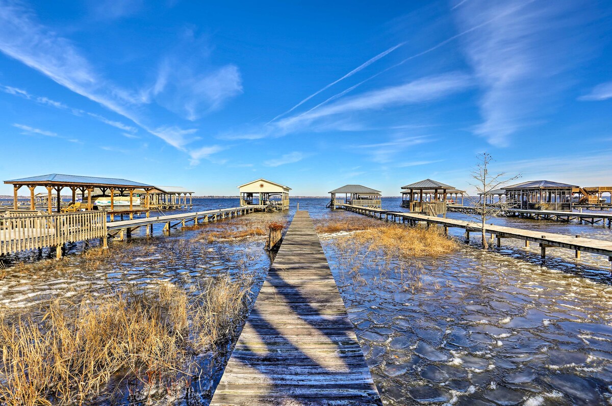 A wooden pier extends over water, leading to several covered structures in the distance. Grassy areas are visible along the edges, with a blue sky and wispy clouds providing a serene backdrop.
