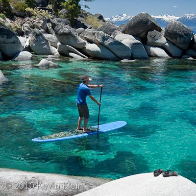 A person is seen paddle boarding on clear turquoise water, surrounded by large rocks and boulders. The backdrop features distant snow-capped mountains under a clear sky. The paddle board appears stable, with the paddler maintaining balance as they navigate the serene water.