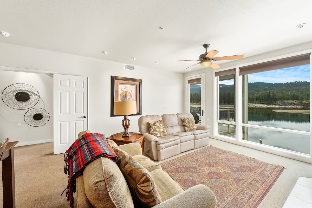 A light-filled living area features comfortable leather seating and a decorative area rug. Large windows provide unobstructed views of the Spokane River and surrounding nature, enhancing the serene ambiance of this inviting space.