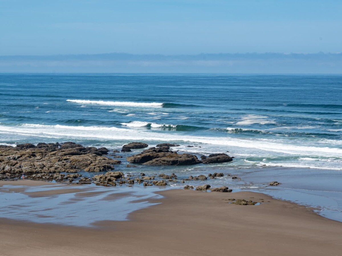 A serene ocean view is presented, featuring gentle waves lapping against rocky formations near the shore. The expansive sandy beach is visible, with a clear blue sky enhancing the coastal scene. The rhythmic flow of the water creates a peaceful ambiance.