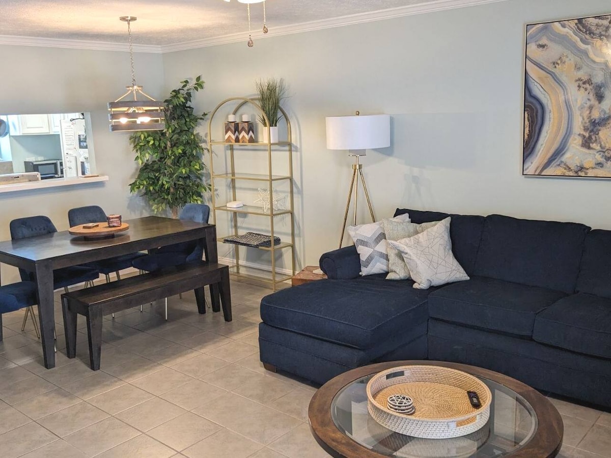 A spacious living area features a large sectional couch in dark fabric, accompanied by a round coffee table. A dining table with blue chairs is visible, and a shelving unit showcases decorative items. Natural light illuminates the space through an open kitchen area in the background.