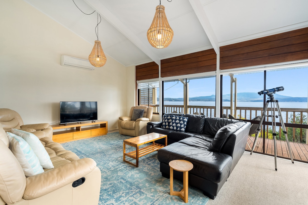 A spacious living area offers a comfortable seating arrangement, featuring two reclining chairs and a sectional sofa. Large windows frame views of the water, while a television and a telescope are positioned for leisure activities. Natural light floods the room, enhancing the serene ambiance.
