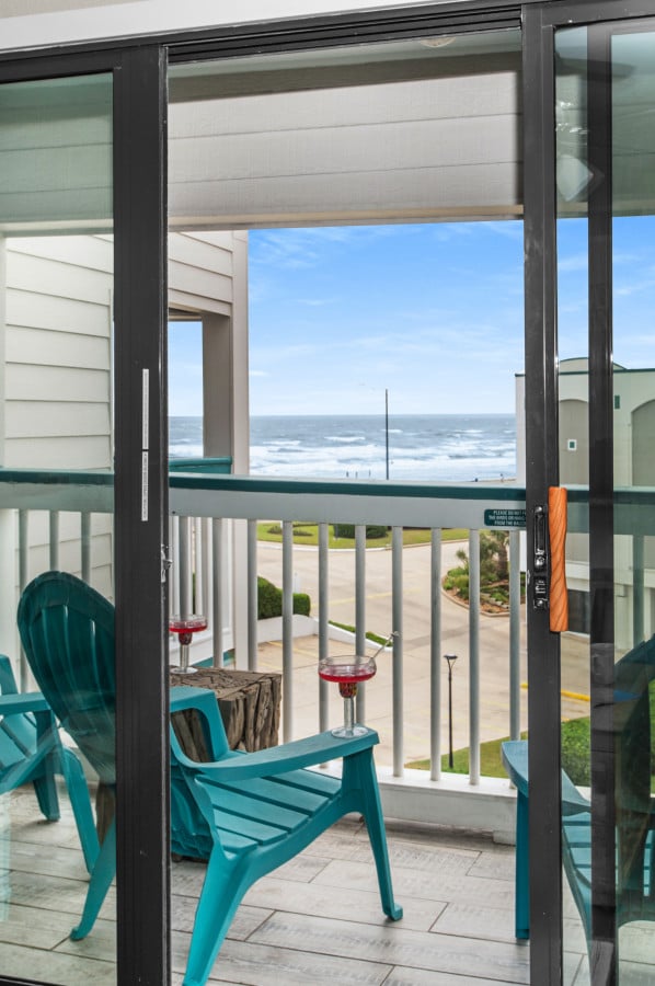 A balcony is visible, featuring two turquoise chairs and a small table. Views of the ocean and coastline can be seen beyond the railing, with waves gently crashing against the shore under a clear sky.