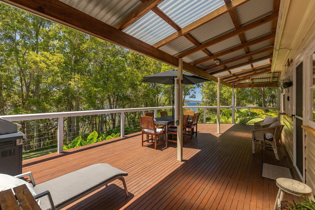 A spacious wooden deck features a dining table and chairs, along with a lounge chair and patio umbrella. A barbecue grill is visible, providing a comfortable outdoor area. Lush greenery surrounds the space, enhancing the connection with nature.