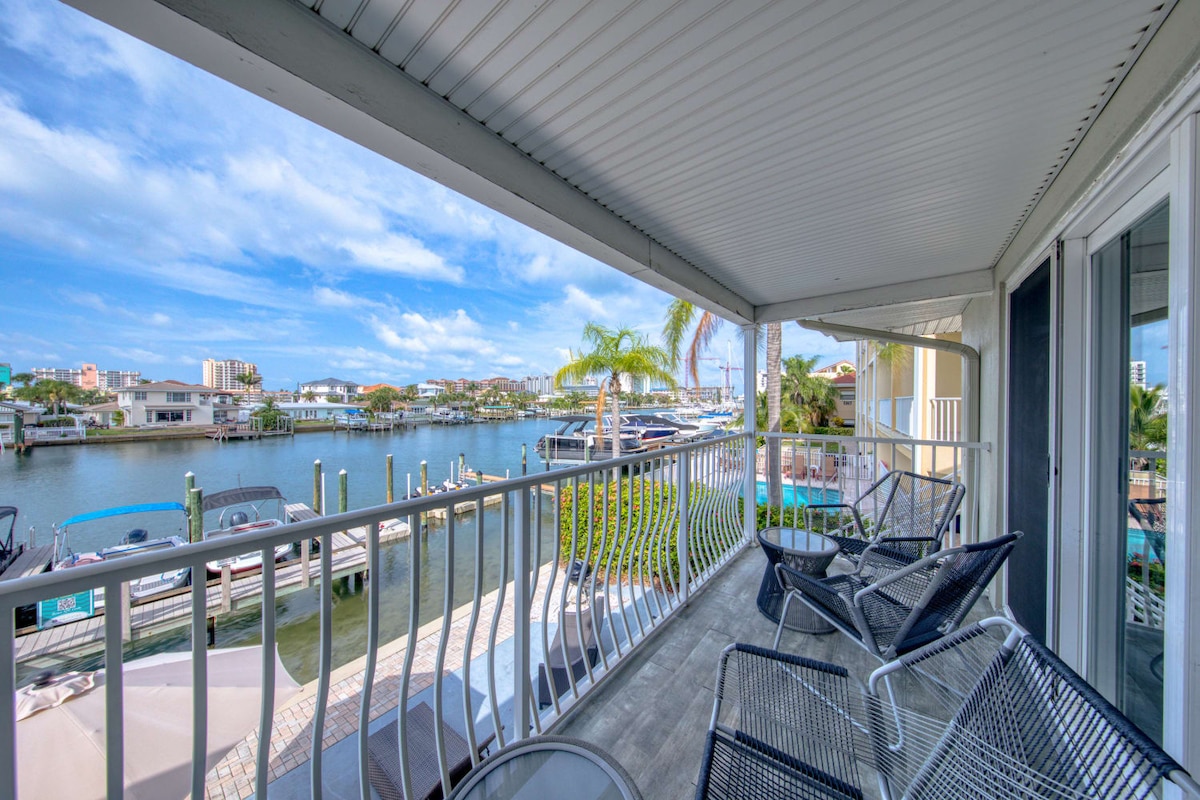 A covered balcony offers a view of the Intracoastal Waterway, framed by palm trees and blue skies. Comfortable chairs are arranged for relaxation, with glimpses of boats docked below and greenery along the riverbank in the background.