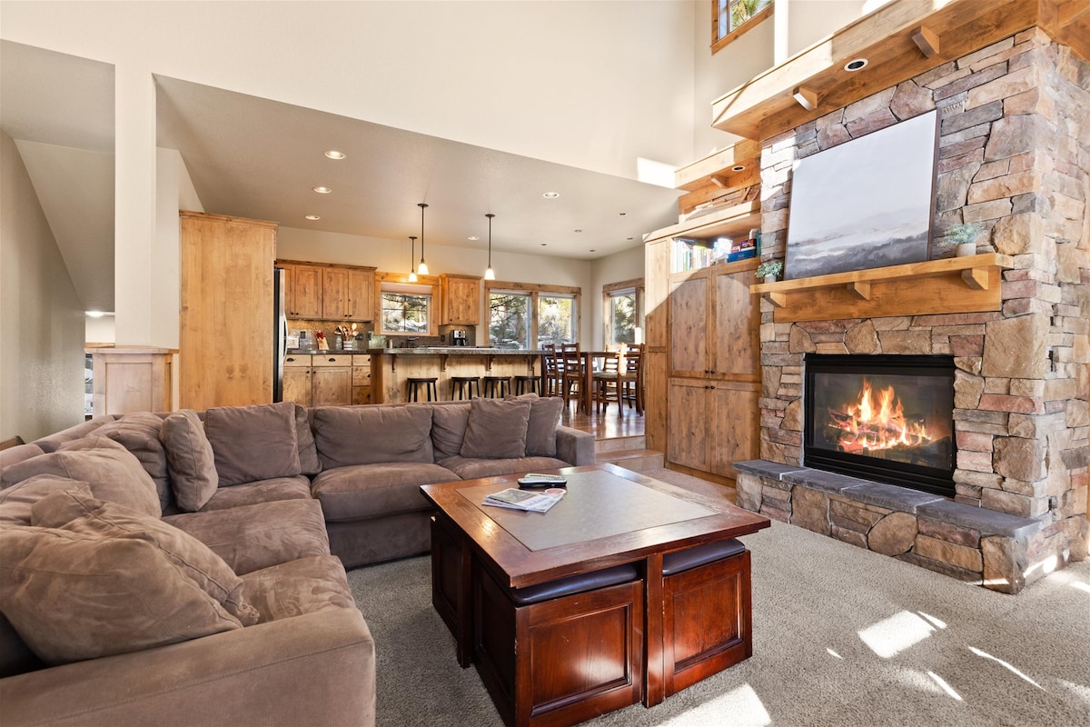 A spacious living room features a large sectional sofa arranged around a central coffee table. A stone fireplace built into the wall provides a cozy ambiance, with light reflecting from a nearby window illuminating the room. The open layout connects to a dining area and kitchen in the background.