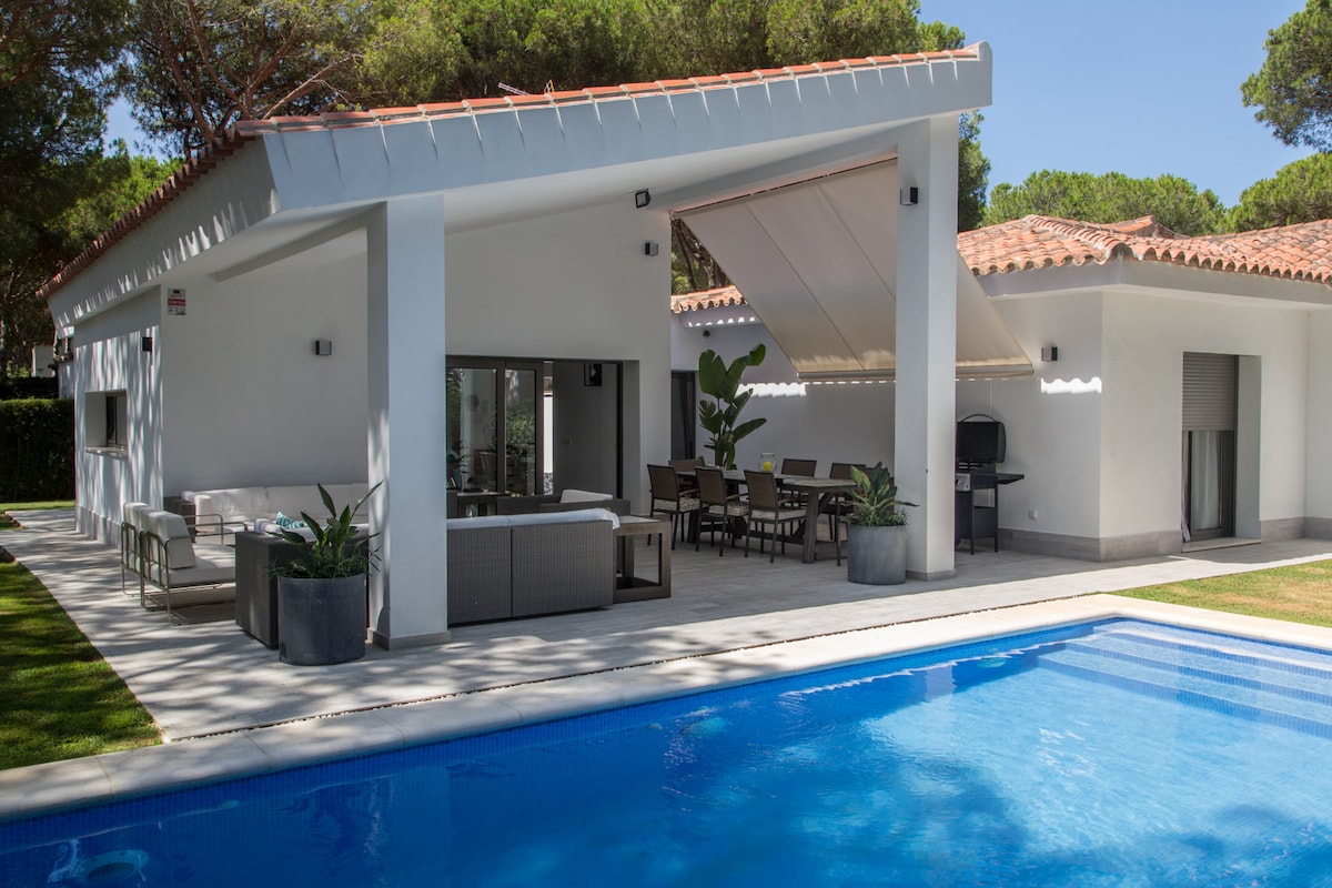 A modern outdoor area is showcased, featuring a swimming pool bordered by a spacious terrace. A covered seating space with comfortable furniture and a dining area is visible, surrounded by lush greenery. The house has large sliding glass doors offering a seamless connection to the poolside.