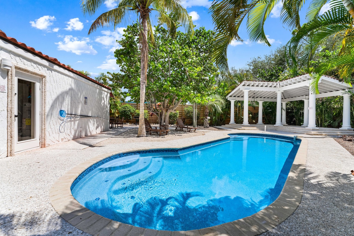 A sparkling pool is surrounded by lush tropical landscaping. The pool area features a spacious patio with lounge chairs, and a pergola stands nearby, providing shaded seating. Bright blue skies enhance the inviting atmosphere of this outdoor oasis.