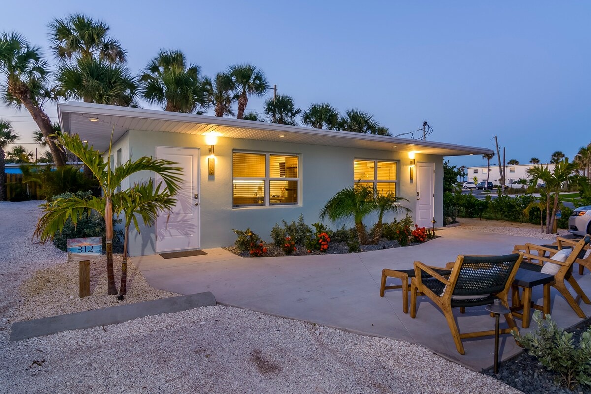 The exterior of the apartment features soft blue walls and modern lighting, enhancing the inviting entrance. Palm trees and vibrant landscaping surround the property. A small seating area with wooden chairs is positioned on the patio, offering a relaxed atmosphere near the entrance.