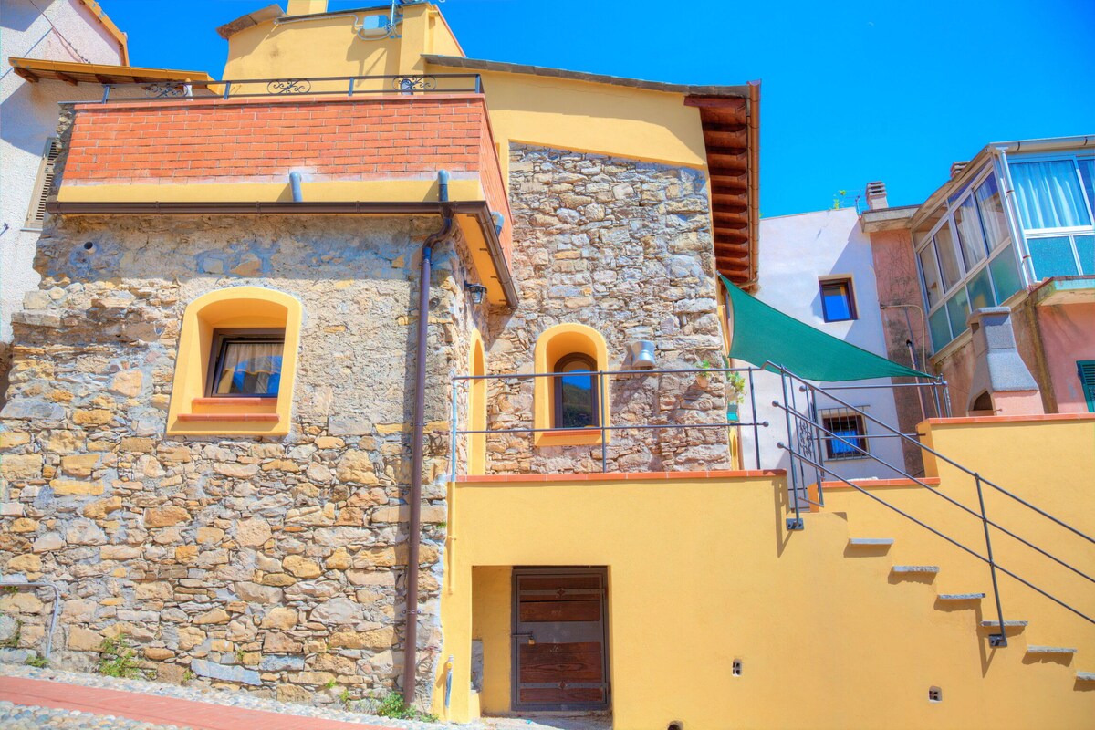 A charming two-story house with stone and yellow plaster exterior is featured. The entrance is framed by brick accents and a staircase leads to the front door. A green awning provides shade over a small outdoor area, enhancing the inviting appearance.