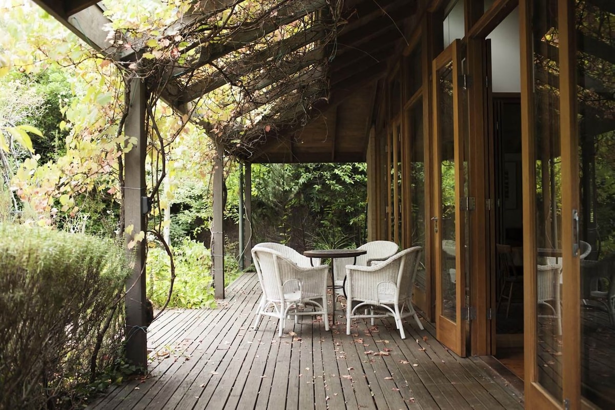 A covered outdoor area features a small round table surrounded by four wicker chairs. Lush greenery expands in the background, and the wooden deck showcases a natural finish. Overhead, a canopy of climbing plants filters the sunlight, providing a serene, shaded space.