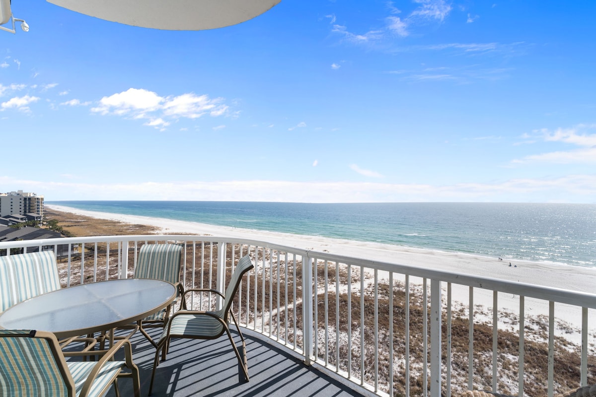 A covered terrace is showcased with outdoor seating for several guests. A glass table sits centrally, surrounded by striped chairs, offering unobstructed views of the pristine beach and the emerald waters of the Gulf Coast under a clear blue sky.