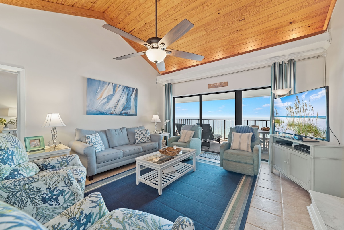 A spacious living area is highlighted by a high, wood-paneled ceiling and a large window offering views of the outdoor landscape. Comfortable seating options, including a light blue sofa and patterned armchairs, surround a central coffee table. A flat-screen TV sits on a light-colored media unit.