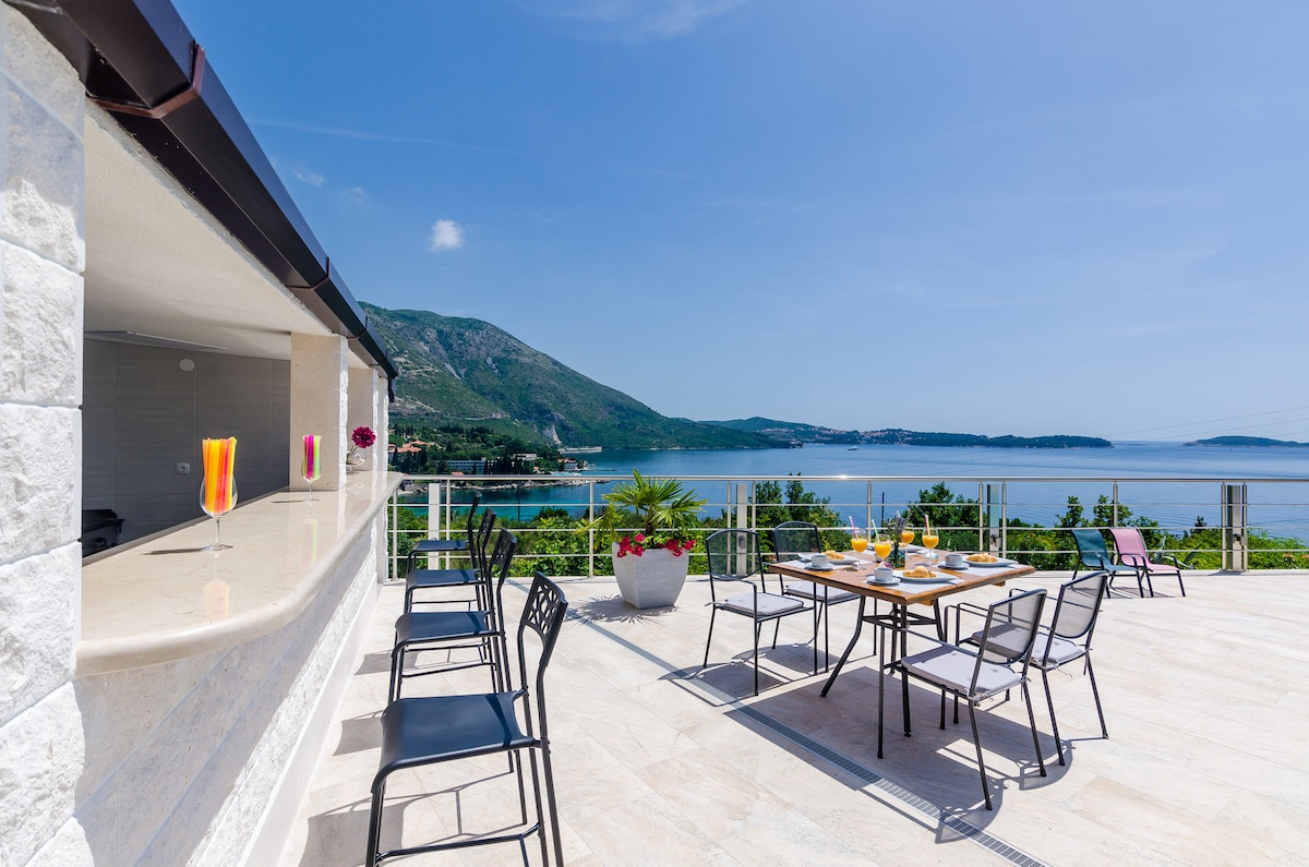 A spacious terrace offers stunning views of the sea and surrounding mountains. Outdoor seating arrangements include several chairs around a table, with a bar area nearby. Bright sunshades add color to the scene, complementing the serene coastal landscape.