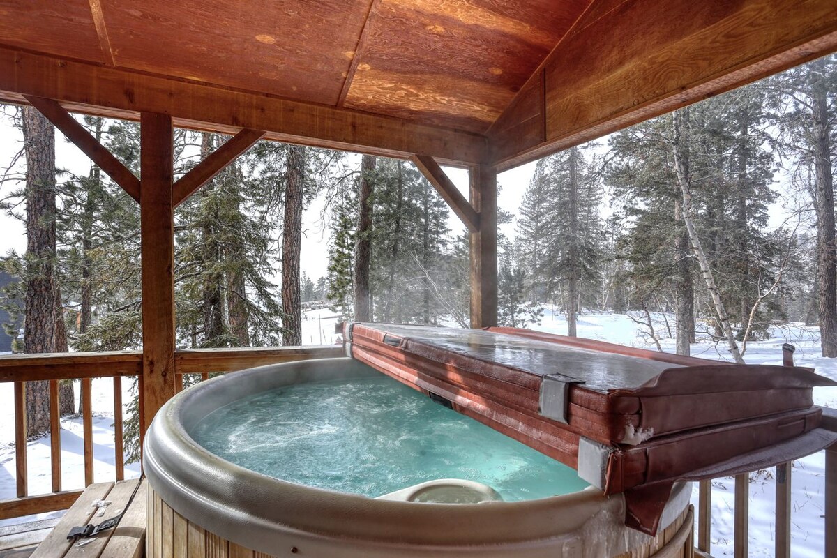 A wooden hot tub is situated on a covered deck, surrounded by a serene winter landscape. Trees are visible in the background, with snow blanketing the ground. A gentle steam rises from the water's surface, creating a calming atmosphere.