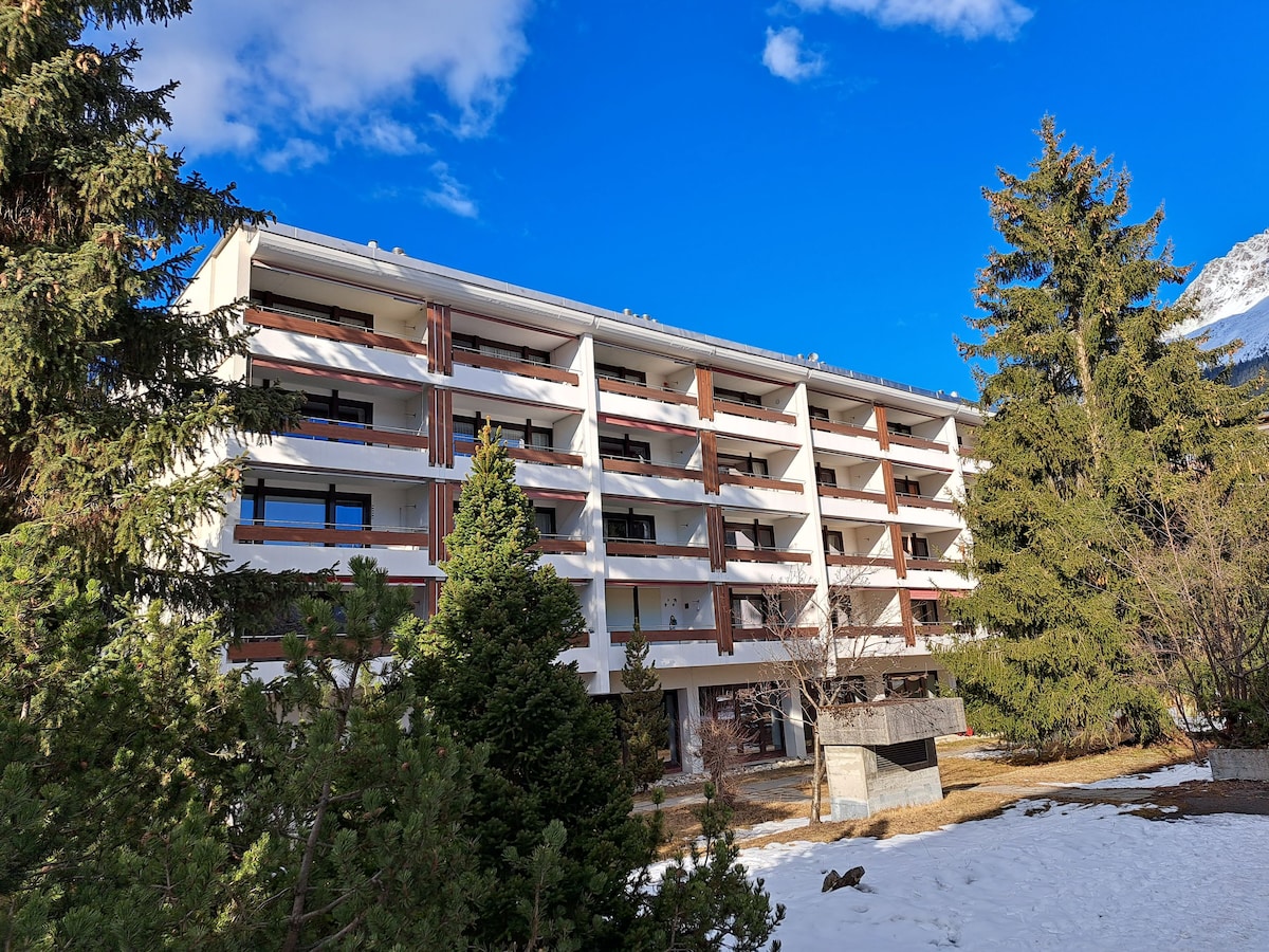 The exterior view of the building presents a four-story structure surrounded by trees and a landscaped area. The design features balconies with railings, and large windows provide natural light. Snow is visible on the ground, indicating the winter season.