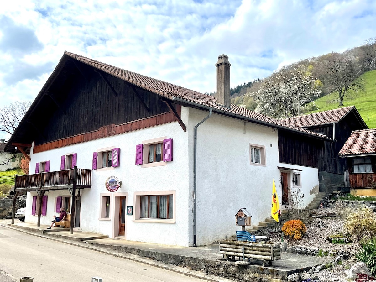 The exterior of the vacation home displays a two-story structure with a combination of white and wooden walls. Purple shutters frame the windows, and a balcony is visible on the upper level. The entrance features a sign, and landscaped stones are arranged in the foreground.