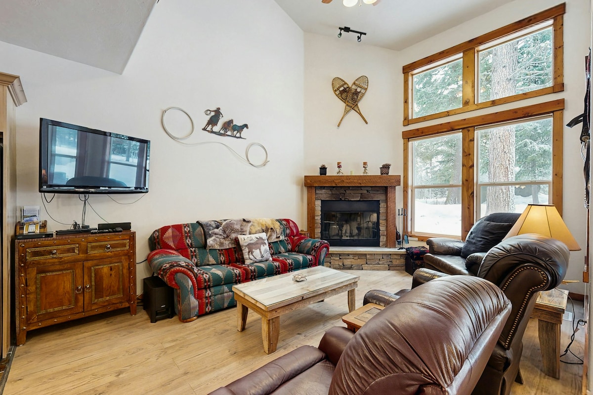 A welcoming living area features a comfortable seating arrangement with a red and green patterned sofa and two recliners. A wooden coffee table sits in front of a stone fireplace, with a television mounted on the wall nearby. Large windows provide views of the surrounding woodland.