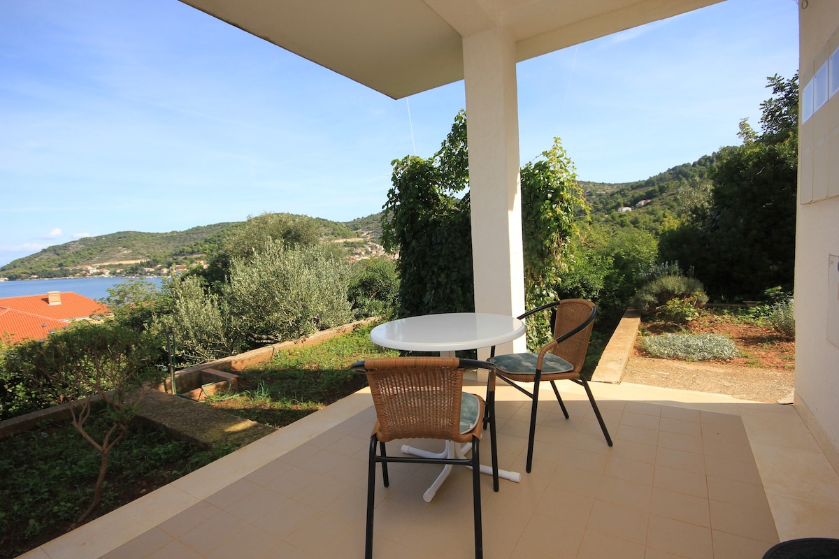 A private terrace features a round table surrounded by two chairs, offering views of the sea and lush greenery. The terrace is shaded by a overhanging roof, enhancing the outdoor space for relaxation.