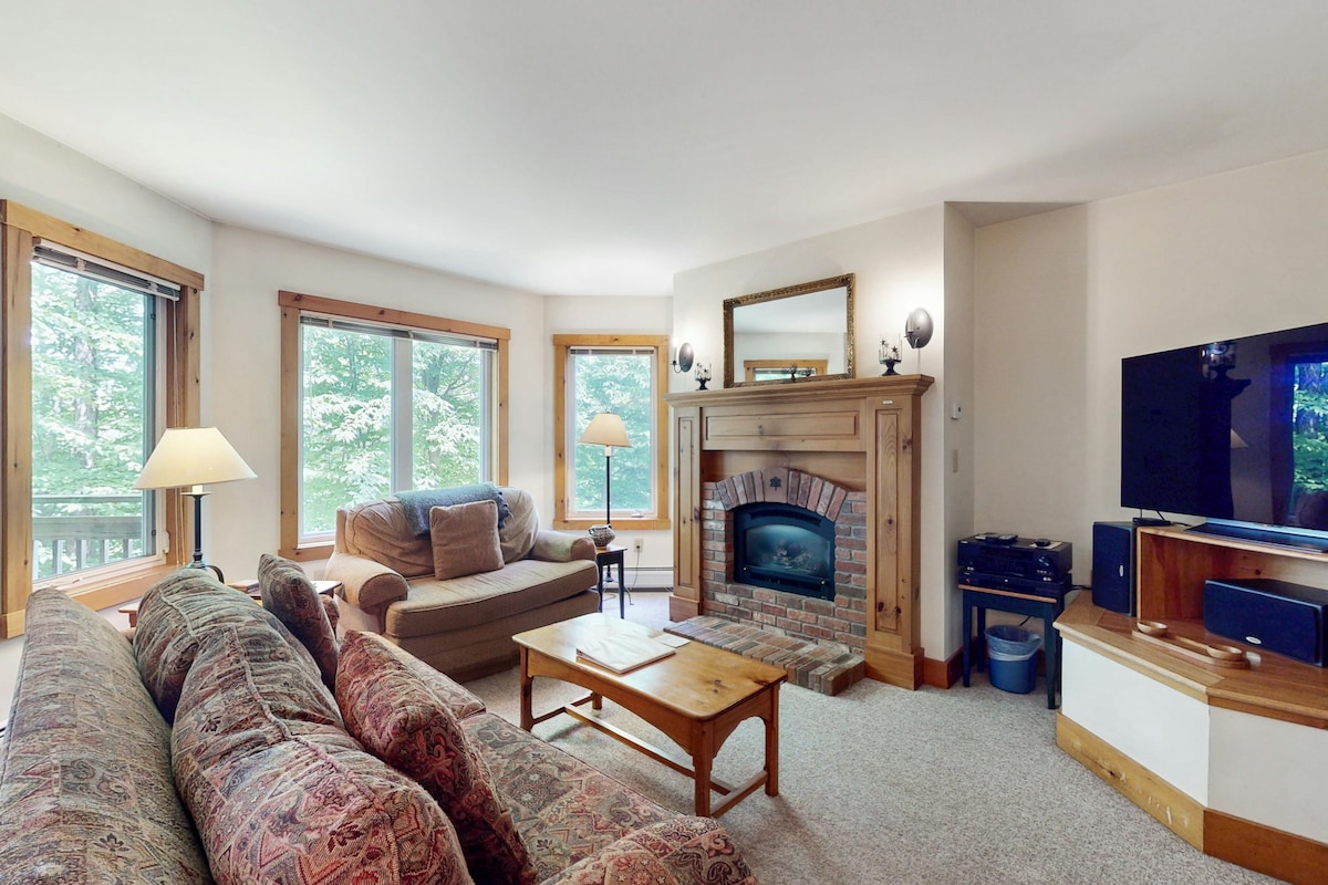 A cozy living area features a gas fireplace with a stone surround, flanked by two large windows offering views of the outdoors. Comfortable seating includes a sofa and an armchair, complemented by a wooden coffee table and an entertainment center with a television.