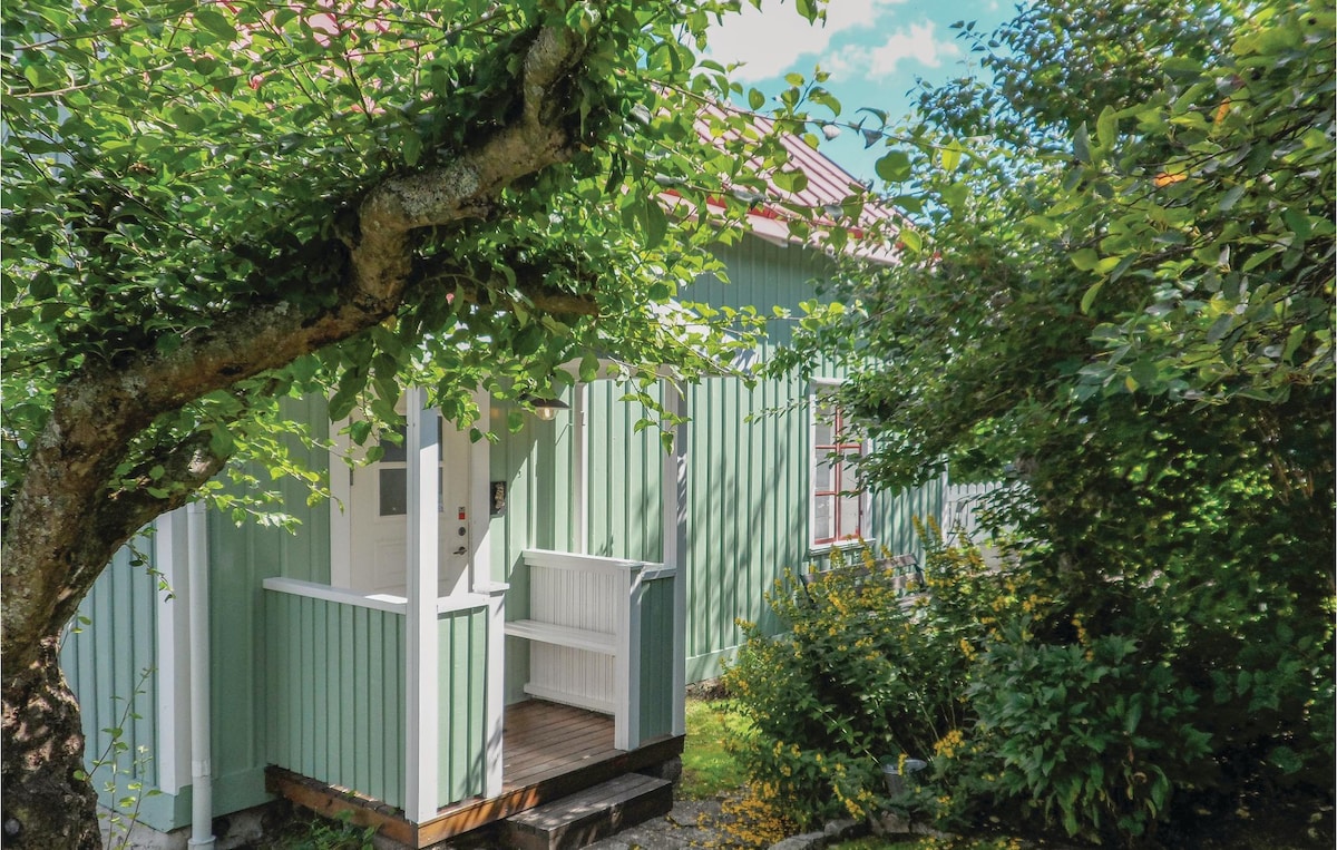 The exterior of a cozy cottage is framed by lush greenery, showcasing a charming light green facade and a welcoming entrance. A tree with broad leaves provides shade, while flowers bloom in the surrounding garden, adding a touch of color to the inviting scene.