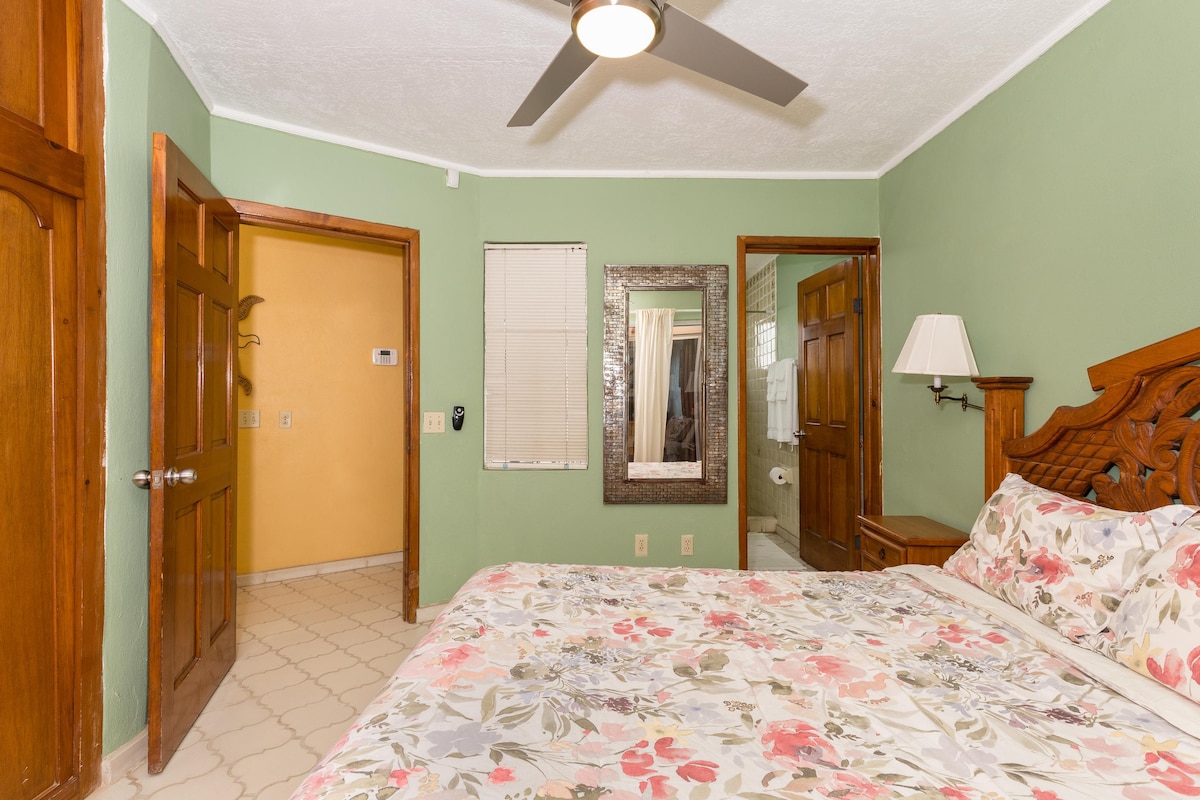 A cozy bedroom features a spacious bed adorned with a floral patterned duvet. Light green walls complement the warm wood furnishings, including a mirror and bedside lamp. The room is illuminated by a ceiling fan and natural light streaming through a nearby window.