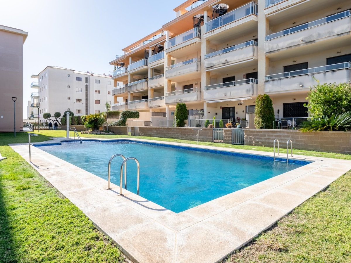 A clear view of a swimming pool surrounded by well-maintained green lawns is presented. The pool is framed by a light-colored concrete border, with ladder access visible. Behind the pool, modern multi-story buildings are seen, featuring balconies and landscaped areas enhancing the overall setting.