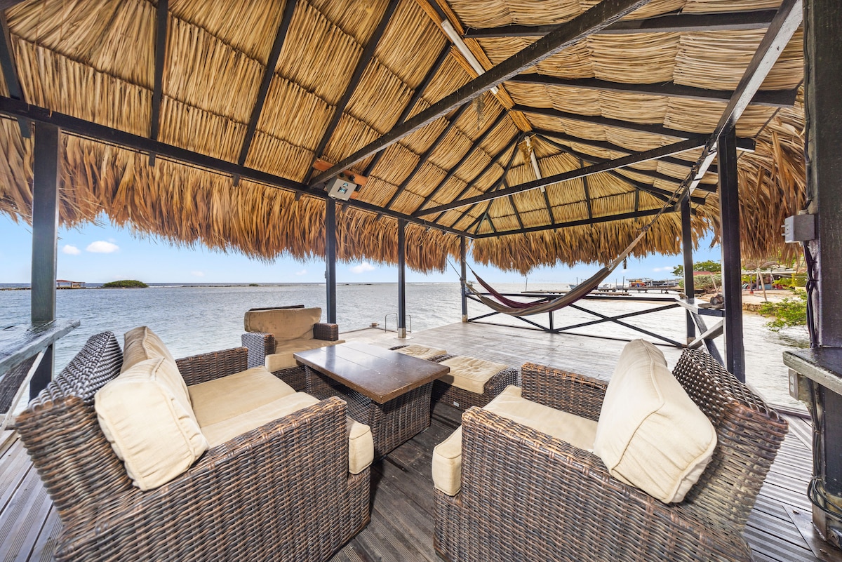 An outdoor seating area beneath a thatched roof is arranged with wicker furniture and plush cushions. A large table is centered, surrounded by comfortable seating, inviting relaxation by the waterfront. A hammock is suspended nearby, offering a cozy spot to unwind while enjoying the scenic views.