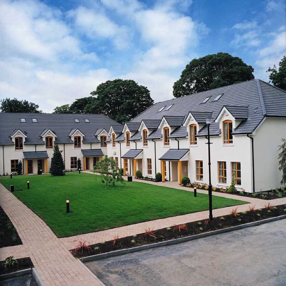 A cluster of modern holiday homes is arranged around a well-maintained courtyard. The properties feature bright yellow window frames, sloped roofs, and are surrounded by landscaped grass, trees, and decorative planting. Pathways made of stone lead to the entrances, creating an inviting atmosphere.
