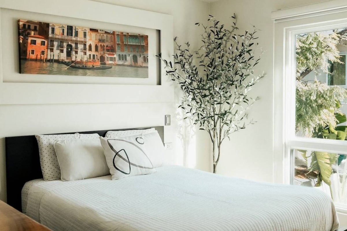 A bright sleeping area features a spacious bed covered with a light-colored blanket. A large, framed artwork depicting a waterfront scene with buildings is mounted above the bed. A decorative tree adds a touch of greenery beside a window allowing natural light to enter.