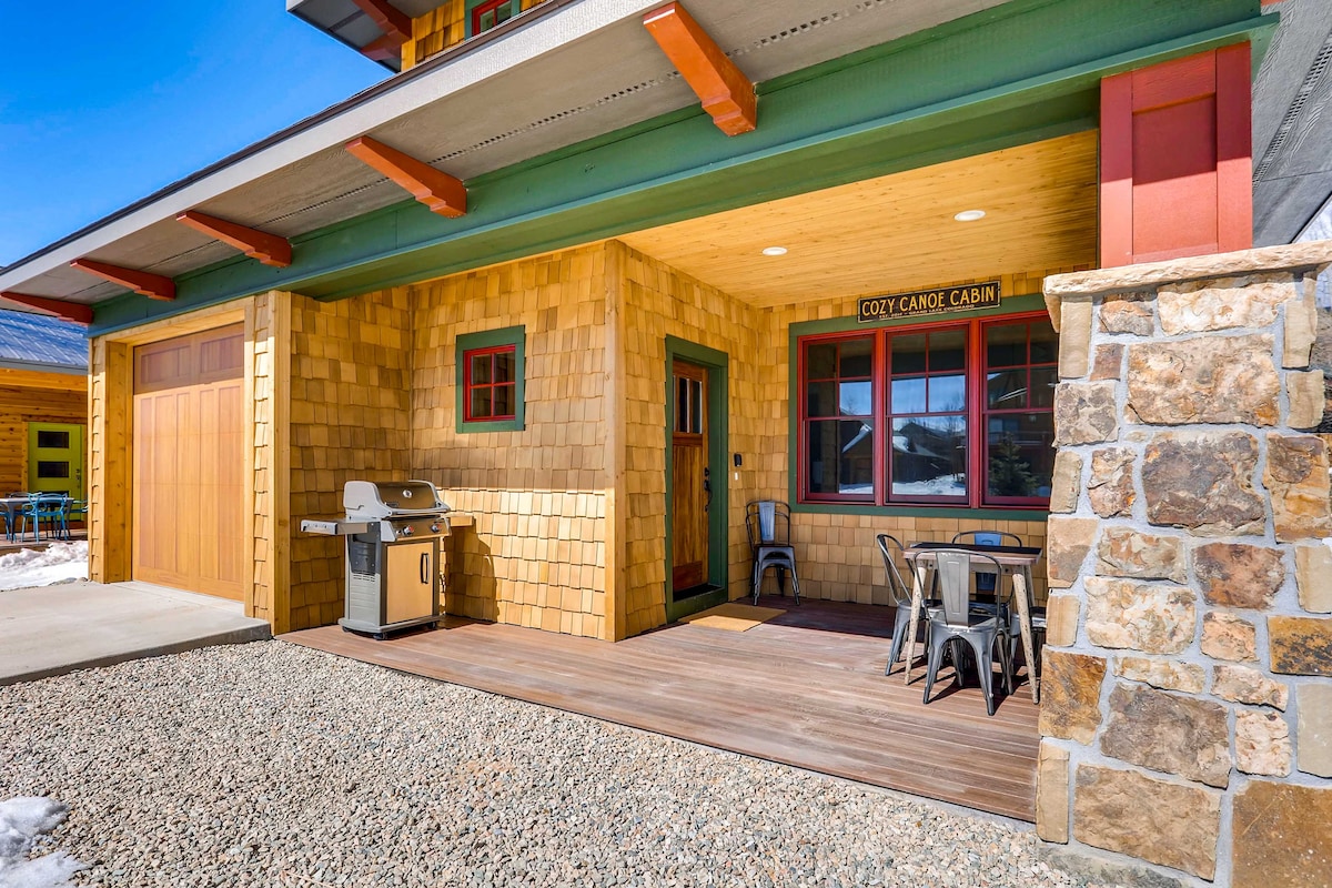 The covered front porch features a dining table with metal chairs, a gas grill, and a textured stone wall. Natural wood accents and large windows are visible, providing a welcoming space with views of the surrounding area. The floor is finished with polished wood planks.