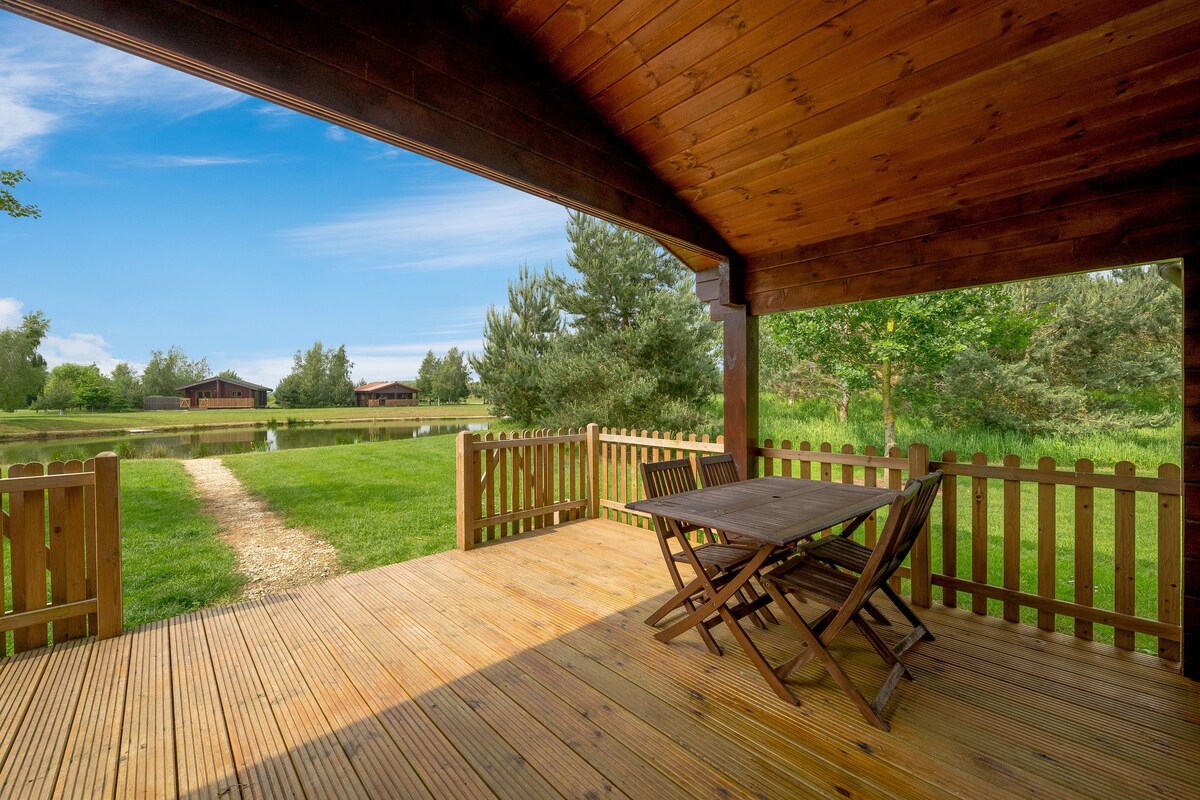 A wooden deck area features a rectangular table surrounded by four folding chairs. The view beyond includes a grassy space leading to a tranquil lake, framed by trees and distant lodges under a clear blue sky.